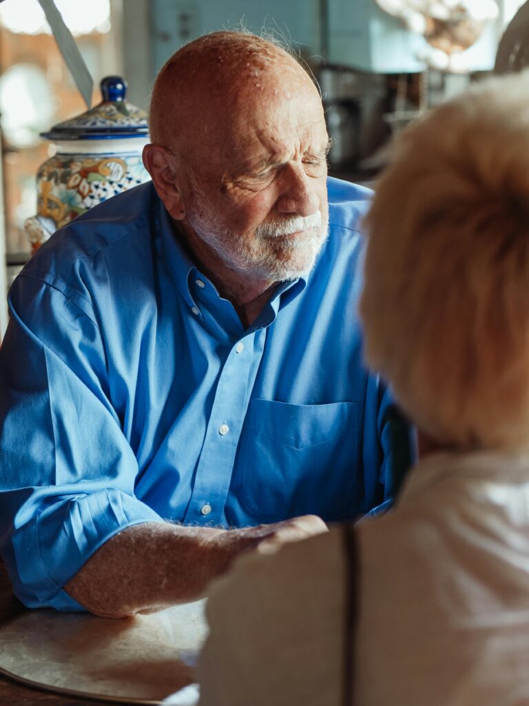 an older man with his eye closed holds hands with someone in the foreground